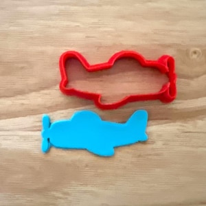 May include: Two airplane-shaped cookie cutters. One is bright red, and the other is a vibrant blue. The cutters are made of plastic and are designed for cutting dough or fondant. The background is a light brown wooden surface.