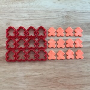 May include: Red plastic teddy bear-shaped cookie cutters are arranged in rows next to orange dough cutouts of the same shape. The items are on a light brown wooden surface.