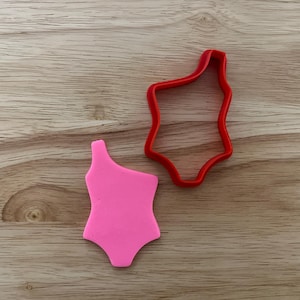 May include: A pink one-shoulder swimsuit and a red swimsuit-shaped cookie cutter sit on a wooden surface. The swimsuit is a solid pink color, and the cookie cutter is a bright red. The items are likely for baking or crafting.