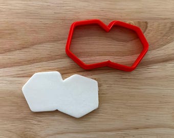 Puede incluir: Un cortador de galletas de plástico rojo con forma de hexágono conectado, junto a una galleta blanca cortada con la misma forma. Los objetos están sobre una superficie de madera.