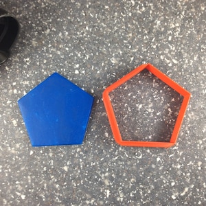 Può includere: Un pezzo blu solido a forma di pentagono e un tagliabiscotti arancione a forma di pentagono. Il pezzo blu è piatto e liscio, mentre il tagliabiscotti arancione ha un centro aperto e pareti sottili. Entrambi sono forme geometriche.