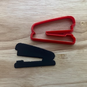 May include: A red plastic cookie cutter in the shape of a stapler sits next to a black stapler-shaped object. The items are on a wooden surface.