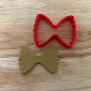 Peut inclure: Un emporte-pièce en plastique rouge en forme de pâtes farfalle à côté d'un biscuit en forme de pâtes farfalle de couleur beige. L'emporte-pièce a un bord festonné. Les pâtes sont sur une surface en bois.
