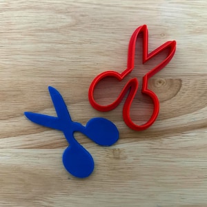 May include: Two plastic scissors-shaped cookie cutters. One is bright red, the other is blue. The cutters are placed on a light brown wooden surface.