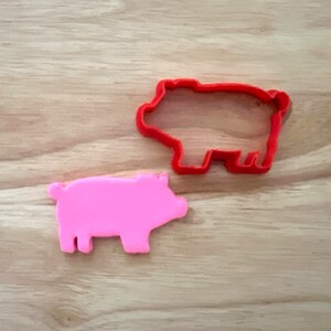 May include: A pink pig-shaped cookie cutter and a red pig-shaped cookie cutter sit on a wooden surface. The pink pig is a solid shape, while the red one is an outline. Both are used for baking.