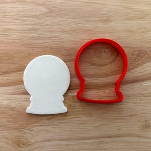 May include: A white, snow globe-shaped cookie cutter and a red snow globe-shaped cookie cutter sit on a wooden surface. The white cookie cutter is a solid shape, while the red one is an outline.