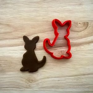 May include: A brown kangaroo-shaped cookie and a red kangaroo-shaped cookie cutter sit on a light wood surface. The cookie is a solid silhouette, while the cutter is a thin outline.