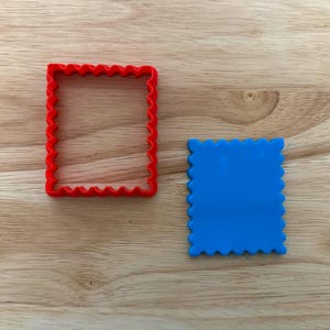 Puede incluir: Un cortador de galletas cuadrado rojo con borde festoneado se encuentra junto a un cuadrado azul con un borde festoneado a juego. Los artículos están sobre una superficie de madera marrón claro.