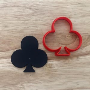 Könnte beinhalten: Ein schwarzer Kleeblatt-Ausstecher und ein roter Kleeblatt-Ausstecher liegen auf einer hellbraunen Holzoberfläche. Das Kleeblattsymbol ist mit Spielkarten verbunden.