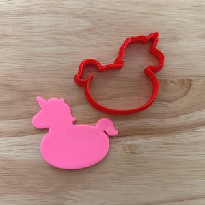 May include: A pink unicorn cookie cutter and a red unicorn cookie cutter sit on a wooden surface. The pink unicorn is a solid shape, while the red one is an outline.