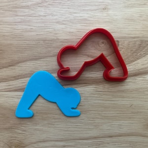 May include: A red and a blue yoga pose cookie cutter on a wooden surface. The red cutter is a solid outline, while the blue one is a filled-in shape. Both are in the downward-facing dog yoga pose.