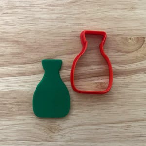 Pode incluir: Um cortador de biscoitos verde em forma de vaso e um cortador de biscoitos vermelho em forma de vaso estão em uma superfície de madeira. O cortador verde é sólido, o vermelho é um contorno. Para assar ou artesanato.