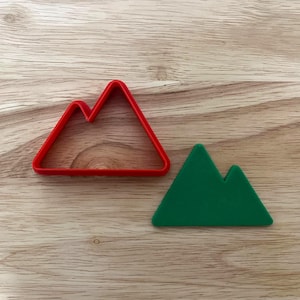 May include: A red plastic mountain-shaped cookie cutter and a green mountain-shaped cookie. The cookie cutter is a simple outline of a mountain range. The green cookie is a solid shape of a mountain range.