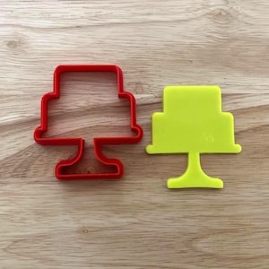 May include: Two cake-shaped cookie cutters. One is a bright red outline, and the other is a solid, vibrant yellow. Both are made of plastic and sit on a wooden surface.