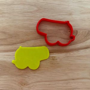 May include: A red plastic cookie cutter and a yellow cookie in the shape of a cannon. The cookie cutter is a simple outline of the cannon shape. The cookie is a solid yellow color and has the same shape as the cutter.