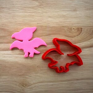 Peut inclure: Un emporte-pièce rose en forme de ptérodactyle et un contour rouge de la même forme sont posés sur une surface en bois. Le biscuit rose est une forme pleine, tandis que le rouge est un contour. Les emporte-pièces sont probablement pour la pâtisserie.
