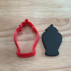 Op de afbeelding: Een rode plastic koekjesvorm en een zwarte koek in de vorm van een decoratieve urn. De koekjesvorm is felrood en de koek is effen zwart. De objecten liggen op een lichtbruin houten oppervlak.