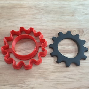 Pode incluir: Dois objetos em forma de engrenagem sobre uma superfície de madeira. Um é um cortador de biscoitos de engrenagem de plástico vermelho brilhante com um centro circular. O outro é um objeto em forma de engrenagem cinza escuro com um centro circular.
