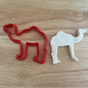 May include: A red plastic camel-shaped cookie cutter next to a white camel-shaped piece of dough. The cookie cutter is a bright red color and has a simple outline of a camel. The dough is white and has been cut into the same shape.