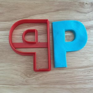 May include: A red plastic letter "P" cookie cutter next to a blue "P" made of dough. The cookie cutter is a single piece with the shape of the letter. The dough is a solid color.