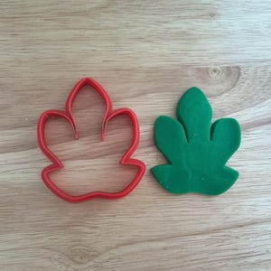 Op de afbeelding: Een rode, blad-vormige koekjessnijder van plastic naast een groen, blad-vormig stuk materiaal. De snijder heeft een gedetailleerde omtrek van de bladvorm. Het groene blad is effen van kleur en lijkt van schuim of vilt te zijn.