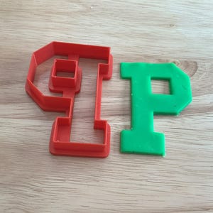 May include: A red plastic letter "P" cookie cutter next to a green letter "P" made of dough. The cookie cutters are on a light brown wooden surface. The cookie cutter is a kitchen tool for baking.