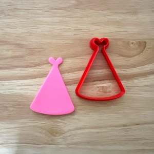 Puede incluir: Dos cortadores de galletas con forma de gorro de fiesta, uno rosa y otro rojo. El rosa es macizo, el rojo es un contorno. Están sobre una superficie de madera clara. Ideales para hornear galletas y decorar pasteles.