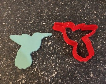 Puede incluir: Dos cortadores de galletas de colibrí, uno rojo y otro azul claro. El cortador rojo está a la derecha y el cortador azul está a la izquierda.