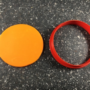 Puede incluir: Un juego de dos cortadores de galletas naranja y rojo. El cortador naranja es un círculo y el cortador rojo es un anillo.