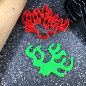 May include: Two cookie cutters, one red and one green, shaped like coral. The cutters are on a grey speckled surface with a wooden rolling pin and black measuring cups.
