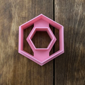May include: Pink plastic cookie cutter shaped like a hexagon with a smaller hexagon cut out in the centre.