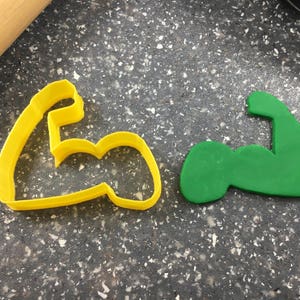 May include: Two cookie cutters in the shape of a bicep, one yellow and one green.