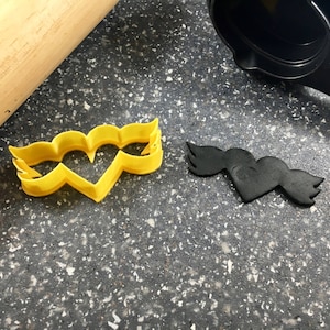 May include: A yellow plastic heart-shaped cookie cutter with wing details, next to a black heart-shaped cookie cutter with wing details. A wooden rolling pin is in the background.