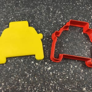 Puede incluir: Dos cortadores de galletas, uno amarillo y otro rojo, con forma de coche. El cortador de galletas amarillo es sólido, mientras que el cortador de galletas rojo es un contorno.