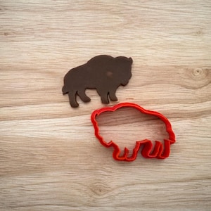 May include: A dark brown bison-shaped cookie cutter and a red bison-shaped cookie cutter sit on a light brown wooden surface. The dark brown bison is a solid shape, while the red cutter is an outline.