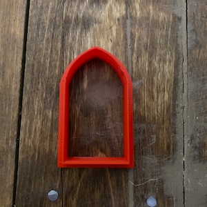 May include: A red plastic cookie cutter shaped like an arched window.