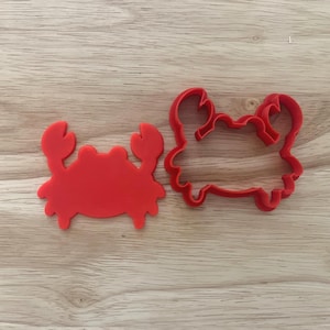 Puede incluir: Dos cortadores de galletas rojos con forma de cangrejo. Uno es una forma sólida de cangrejo, y el otro es un contorno de cangrejo. Los cortadores son de material flexible y están sobre una superficie de madera marrón claro.