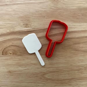 Könnte beinhalten: Zwei Ausstecher in Form eines Eis am Stiel. Einer ist weiß und der andere rot. Die Ausstecher liegen auf einer Holzoberfläche.