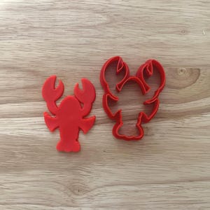 May include: Two red lobster-shaped cookie cutters on a wooden surface. One cutter is a solid lobster shape, and the other is an outline of a lobster. The cutters are made of plastic and are likely used for baking or crafting.