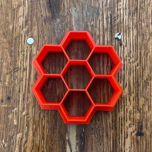 May include: A bright orange, honeycomb-shaped cookie cutter made of plastic. The cutter features a series of interconnected hexagonal cells, perfect for creating bee-themed treats. The cutter is shown on a rustic, wooden surface.