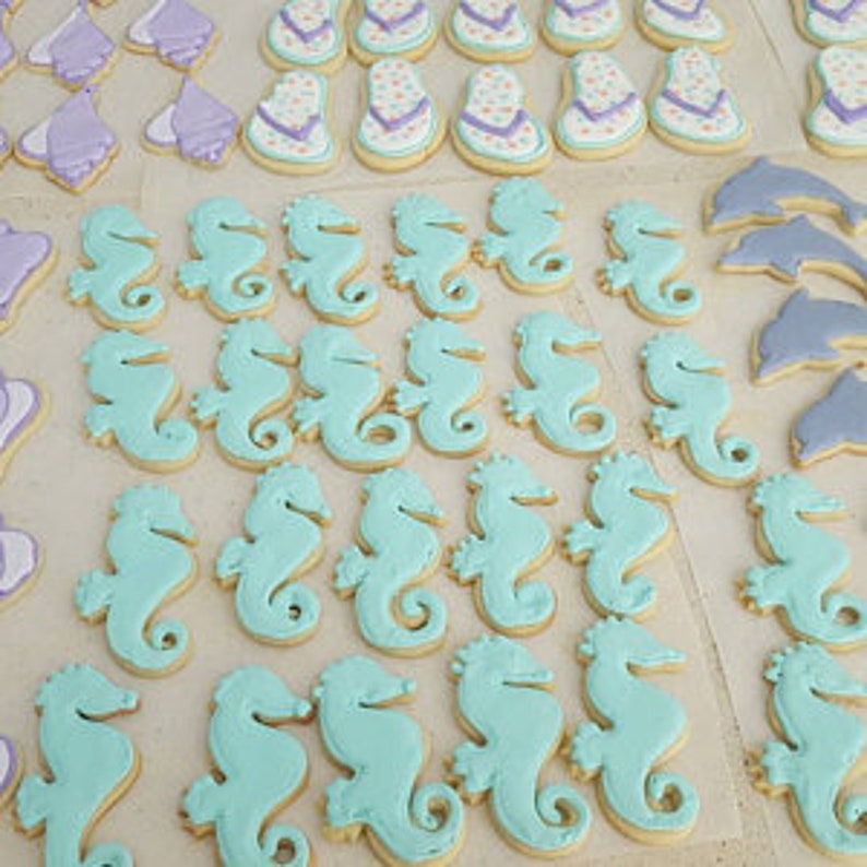 May include: Assortment of decorated sugar cookies in various shapes, including seahorses, dolphins, and wedding cakes. The cookies are decorated with royal icing in shades of blue, purple, and white.