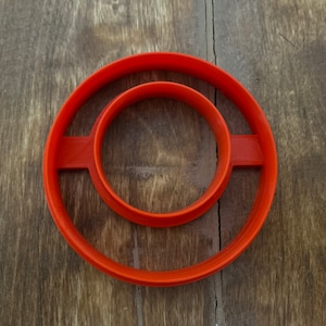 May include: Orange plastic cookie cutter with two concentric circles. The outer circle is larger than the inner circle.