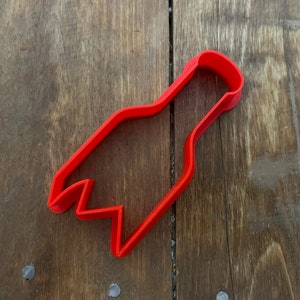 May include: A bright red cookie cutter in the shape of a bottle opener. The cutter is made of plastic and has a handle and a jagged edge for cutting. The cookie cutter is on a wooden surface.