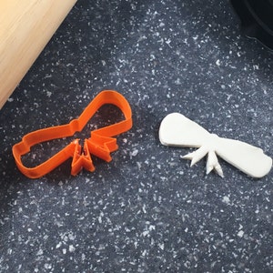 May include: Two plastic cookie cutters, one orange and one white, shaped like bows. The cutters are on a dark gray surface.