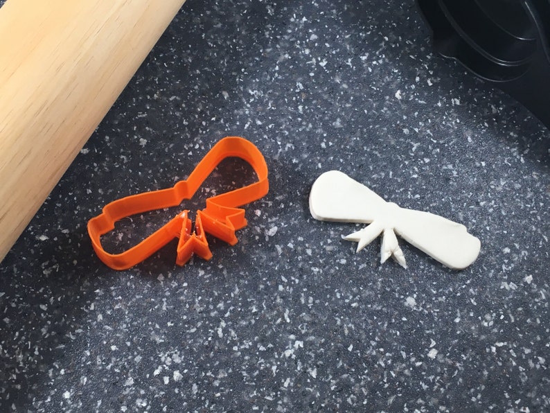 May include: Two plastic cookie cutters, one orange and one white, shaped like bows. The cutters are on a dark gray surface.