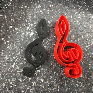 Puede incluir: Dos cortadores de galletas en forma de clave de sol, uno negro y otro rojo, sobre una superficie gris moteada.