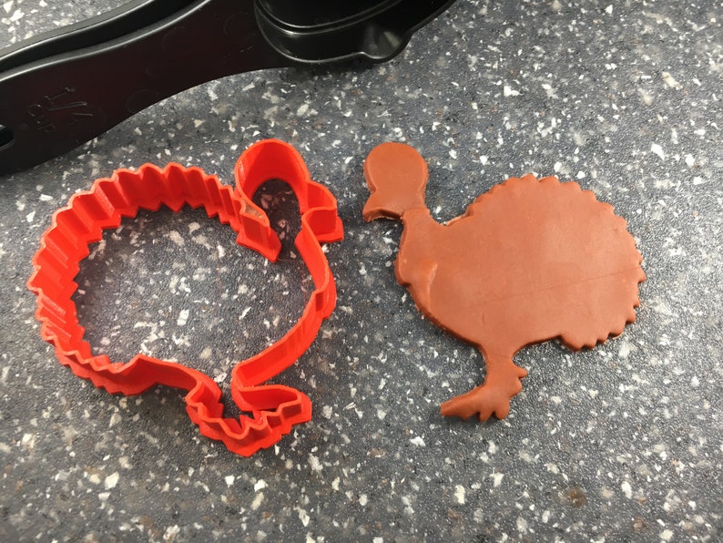 May include: A red plastic turkey cookie cutter and a brown turkey-shaped cookie on a gray countertop.