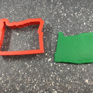 May include: A red plastic cookie cutter shaped like the state of Oregon, next to a green play dough cutout of the state of Oregon.