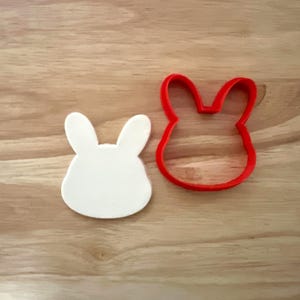 May include: A white bunny-shaped cookie cutter and a red bunny-shaped cookie cutter are displayed on a wooden surface. The white bunny is a solid shape, while the red one is an outline. The image is well-lit and suitable for baking-related content.