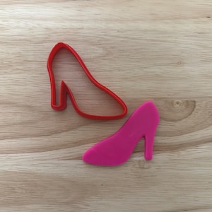 May include: A red outline and a pink high-heeled shoe cookie cutter on a wooden surface. The cookie cutter is in the shape of a high-heeled shoe. The pink shoe is next to the red outline.